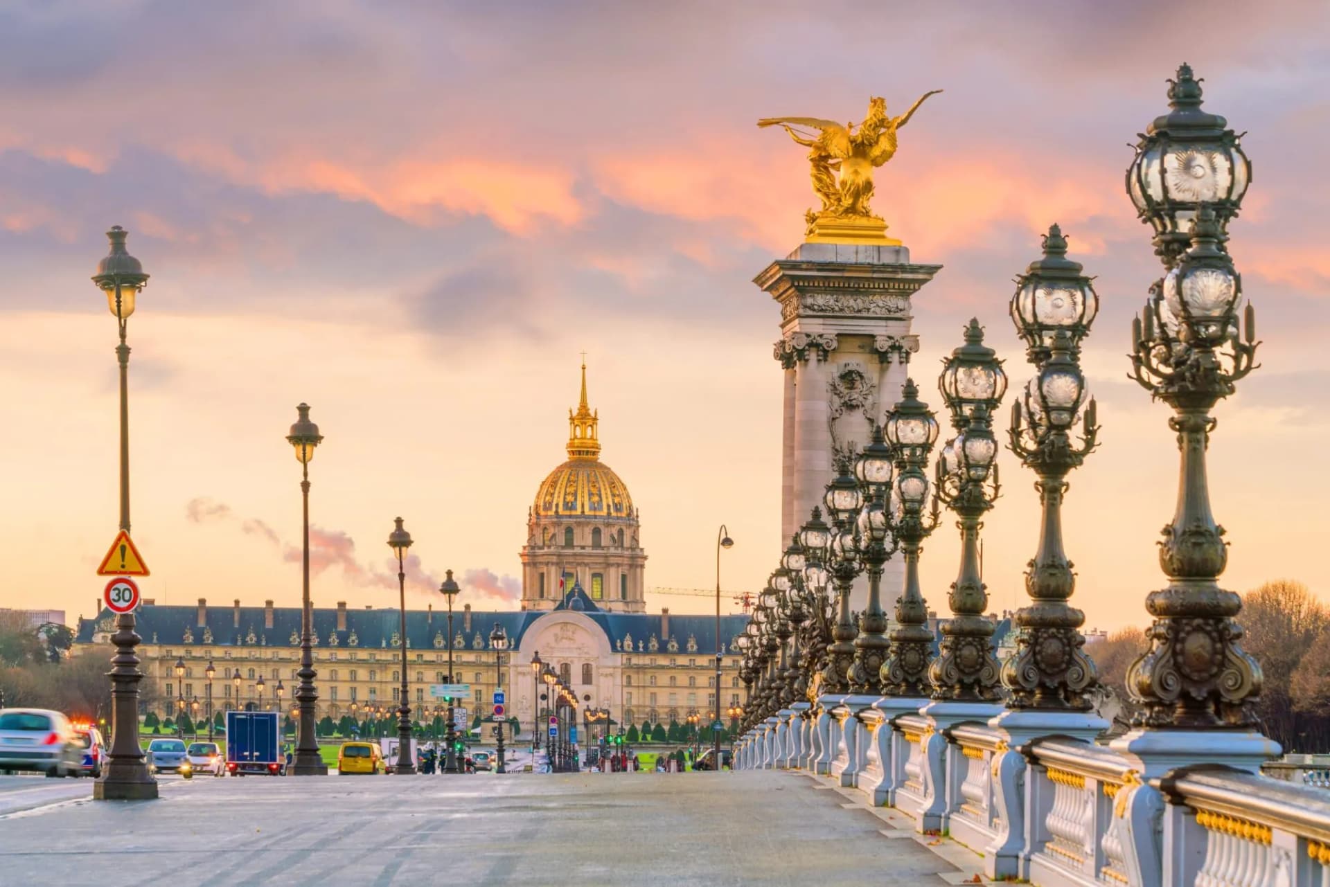 Pont Alexandre III ornate lampposts with Les Invalides dome at sunset in Paris.