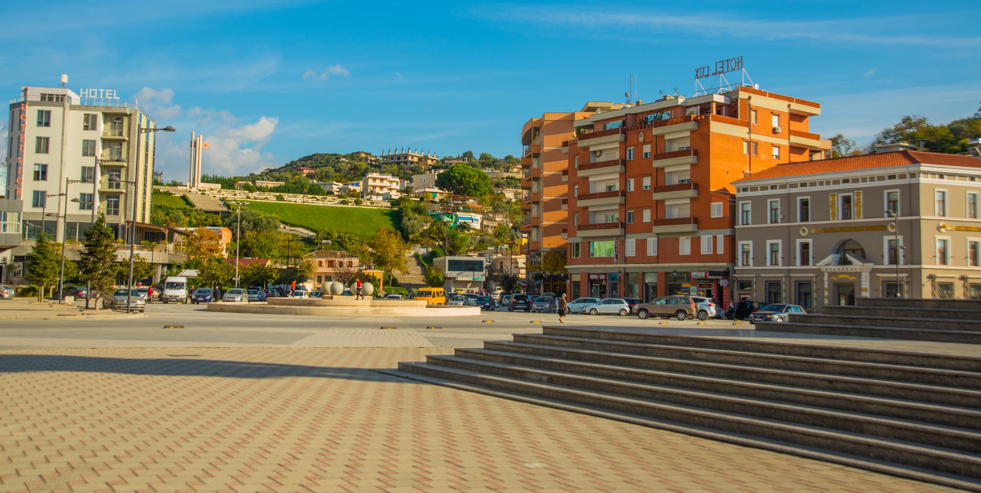 City square with modern buildings, parked cars, and a green hill in Vlore, Albania.