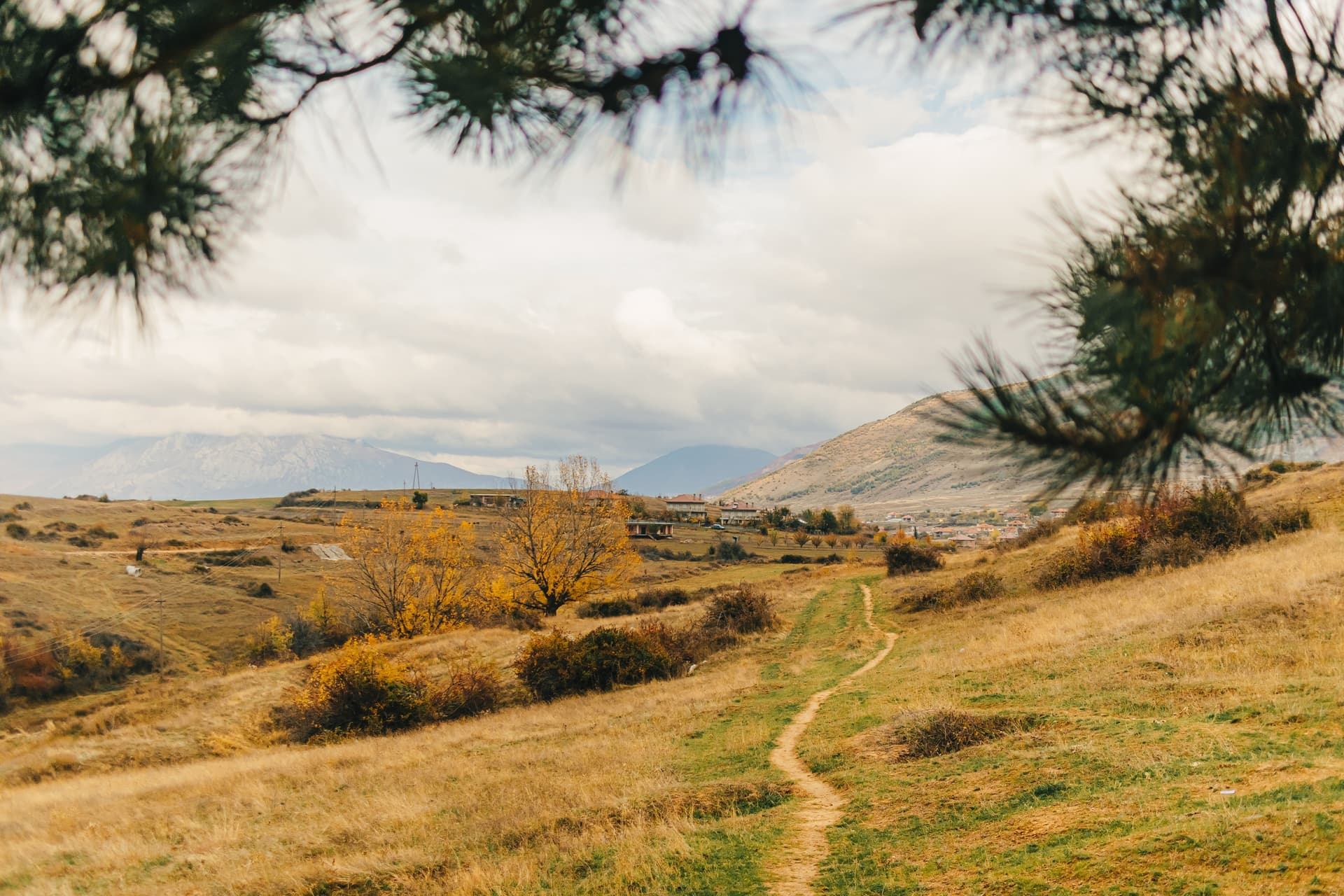 Winding dirt path through dry hills toward village and hazy mountains in Albanian hinterland.