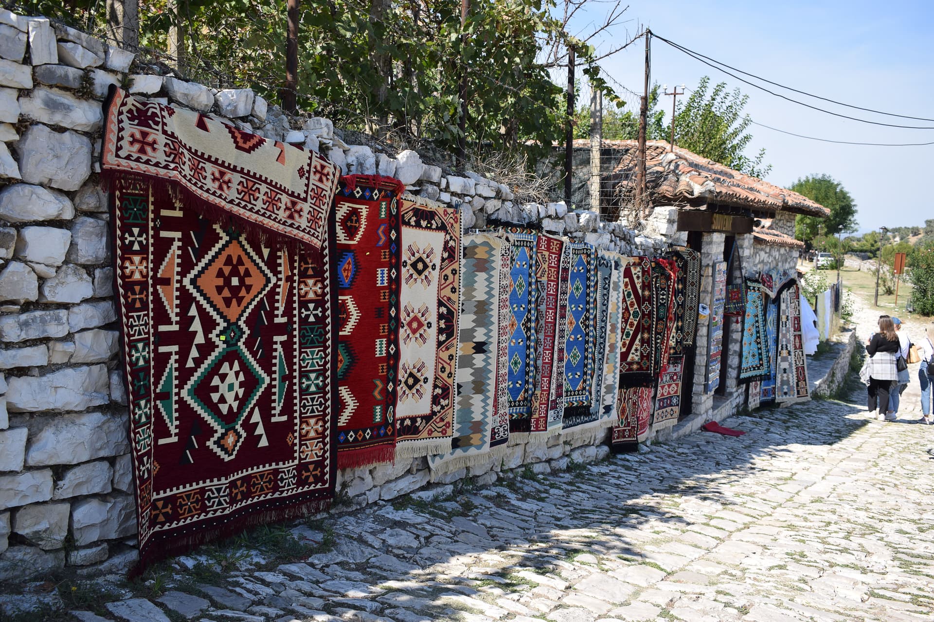 Colorful rugs displayed on a stone wall along a cobblestone path in an Albanian village.