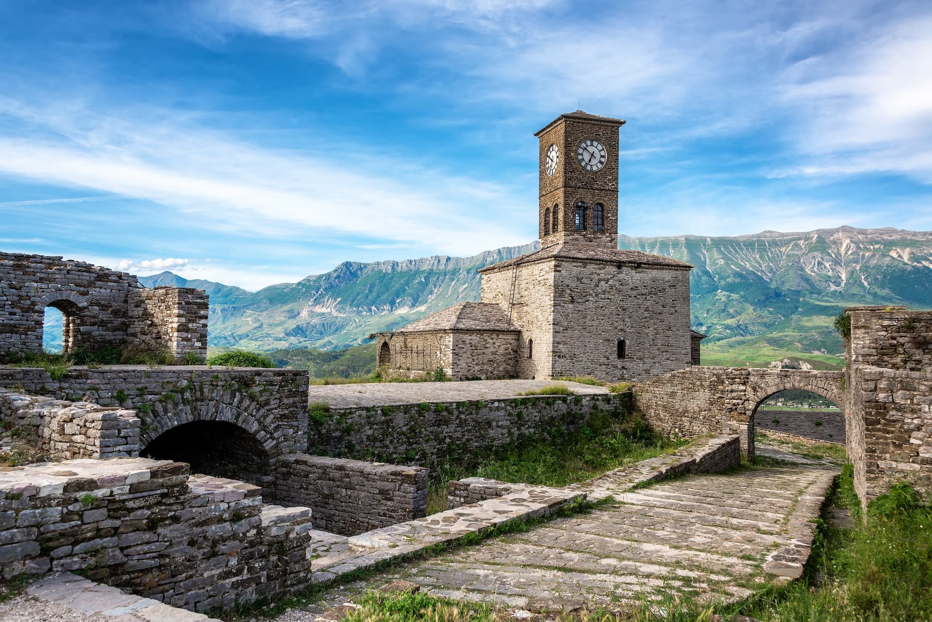 Stone clock tower ruins in Gjirokaster, Albania, with green mountains under a blue sky.
