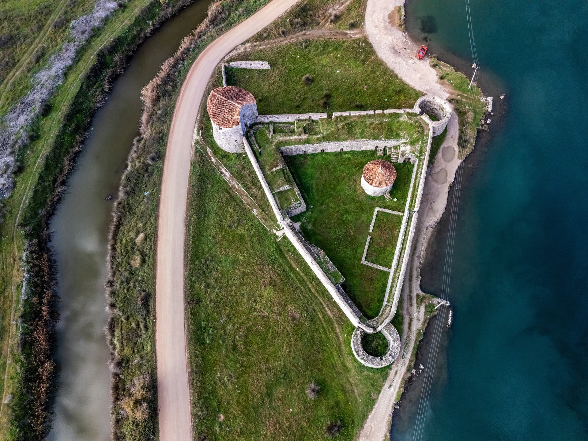 Aerial view of Butrint fortress ruins with stone walls, round towers, and green grass next to a canal and blue water.