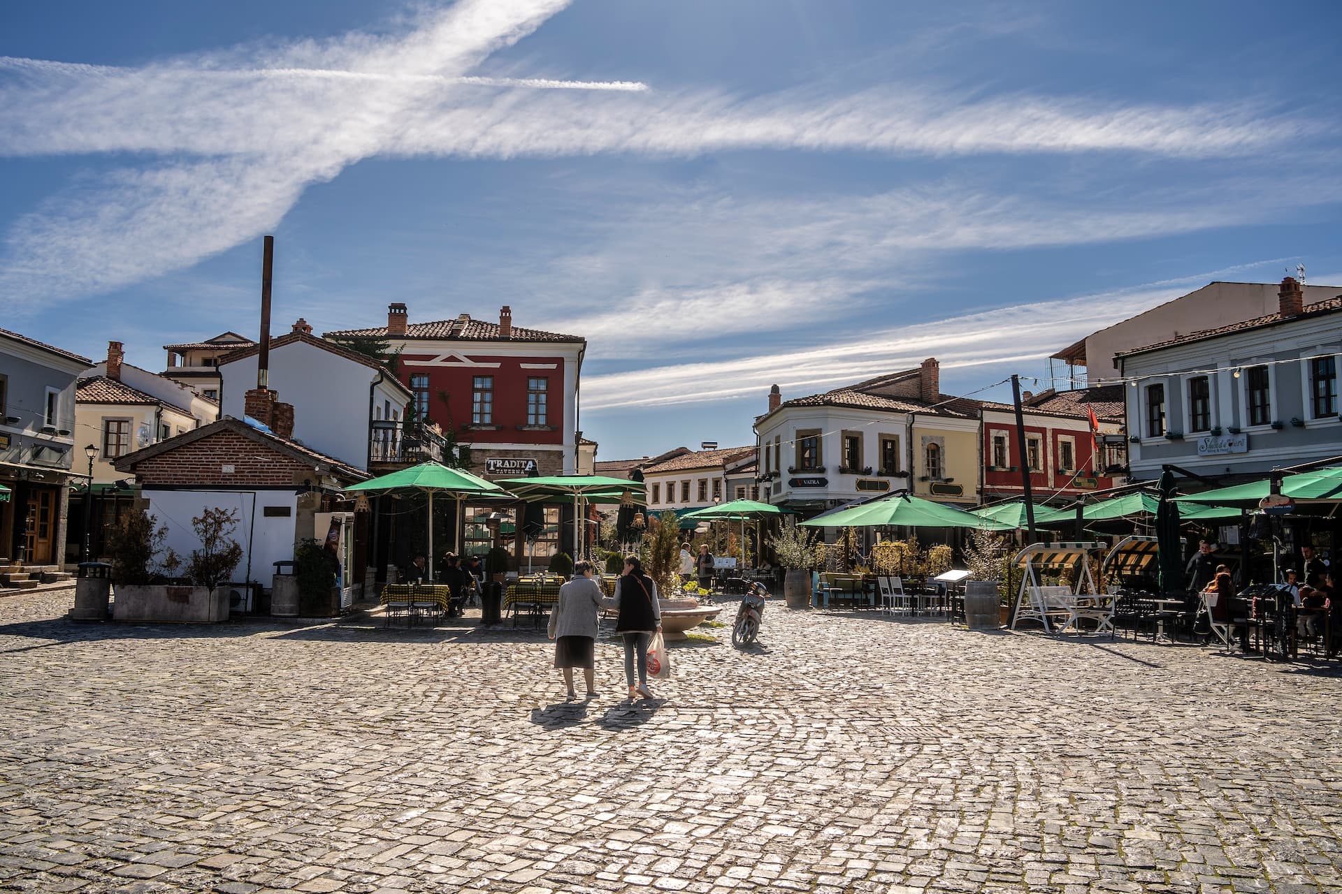 Cobblestone square in Korca, Albania with traditional buildings and outdoor cafe seating under green umbrellas.