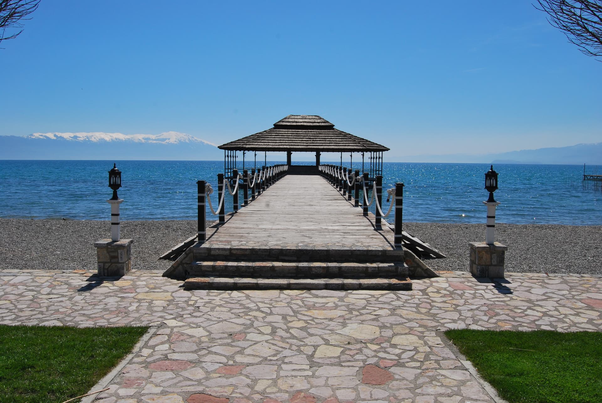 Wooden pier with thatched roof leading to blue water, snow-capped mountains in background at Beach Struga.