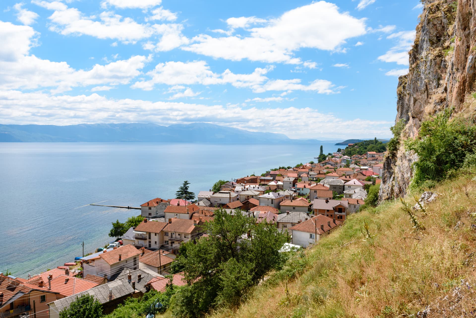 Houses with red roofs line the shore of Lake Ohrid, Macedonia, beneath a steep cliff.