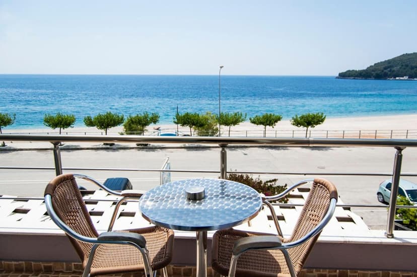 Balcony with wicker chairs and metal table overlooking beach, road, and blue sea in Himare.