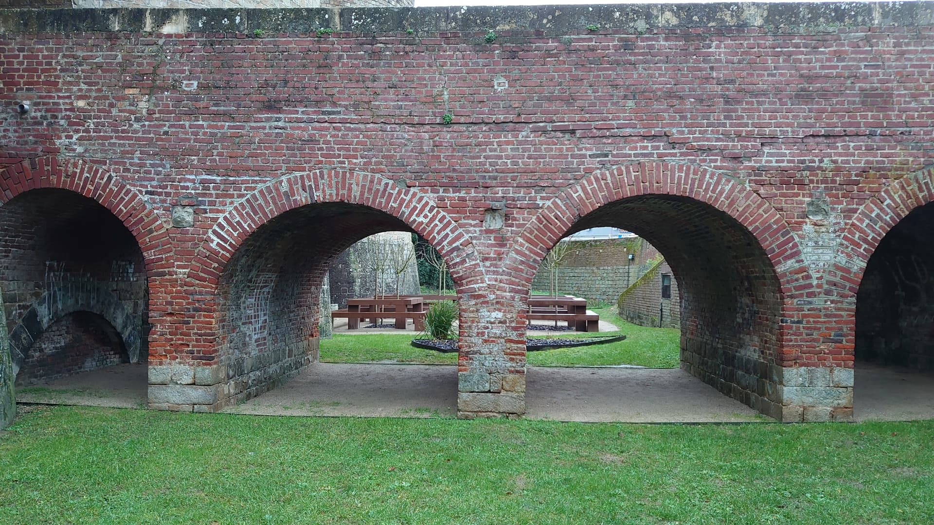 Arched brick wall overlooking a grassy area with wooden picnic tables in Péronne.