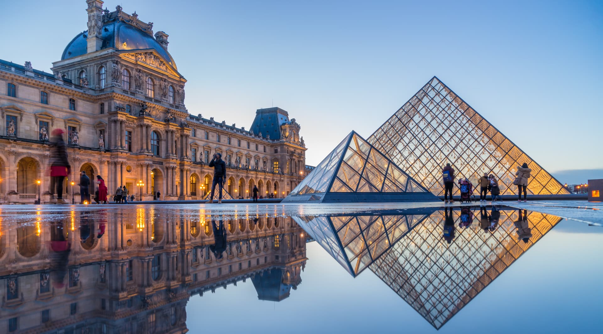 Louvre Pyramid and palace illuminated at dusk with reflections on wet pavement in Paris.