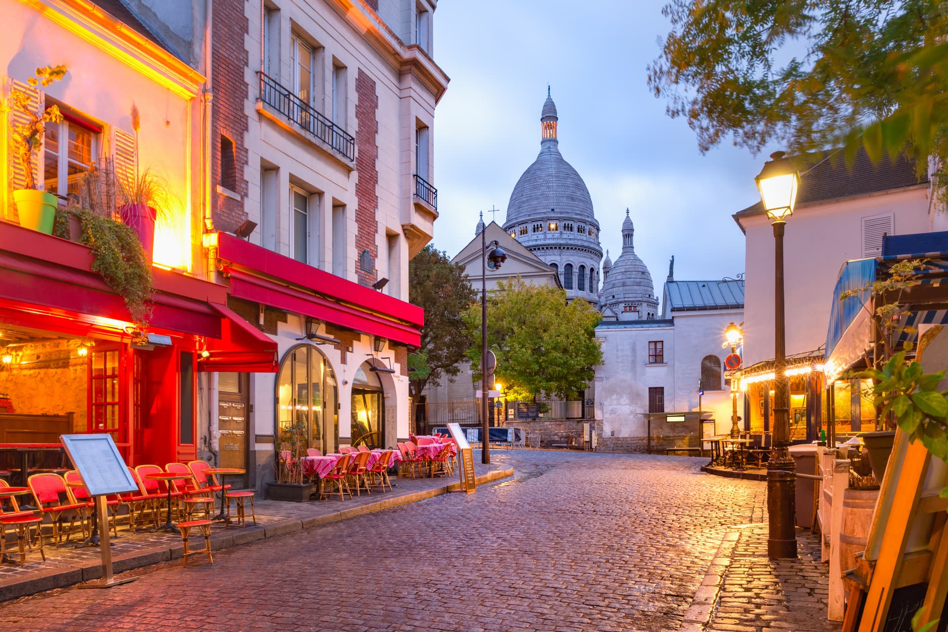 Cobblestone street cafe with red chairs outside, Sacré-Cœur Basilica dome visible in Paris.