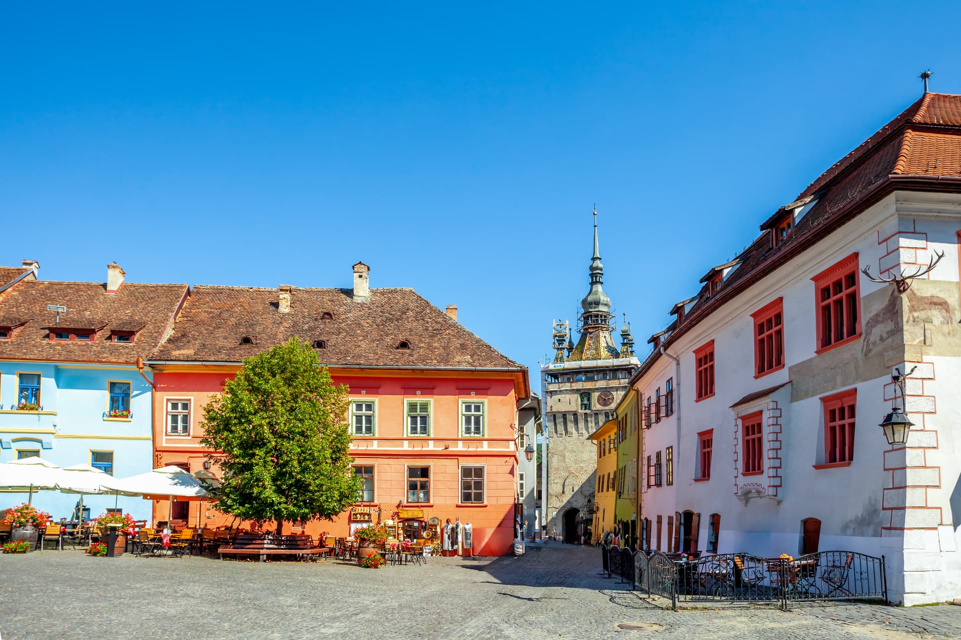 Colorful historic buildings surround Sighisoara Clock Tower in a cobblestone square with outdoor cafe seating.