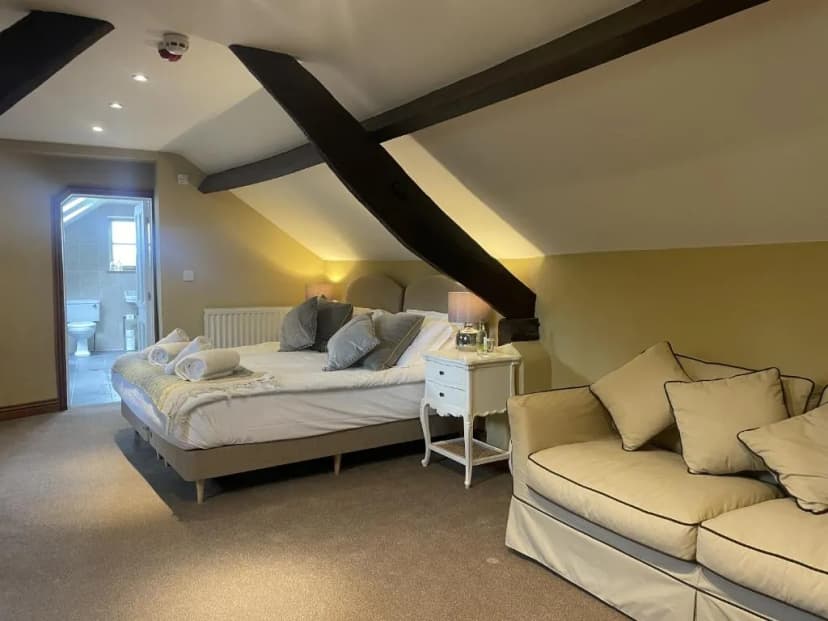 Cozy attic bedroom with exposed dark beams, large bed, sofa, and ensuite bathroom visible.