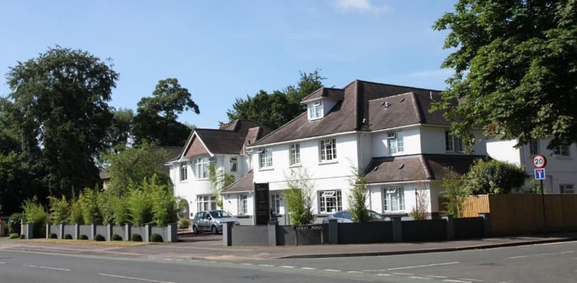 White building with dark roof next to road, with green trees and Apley Road sign.