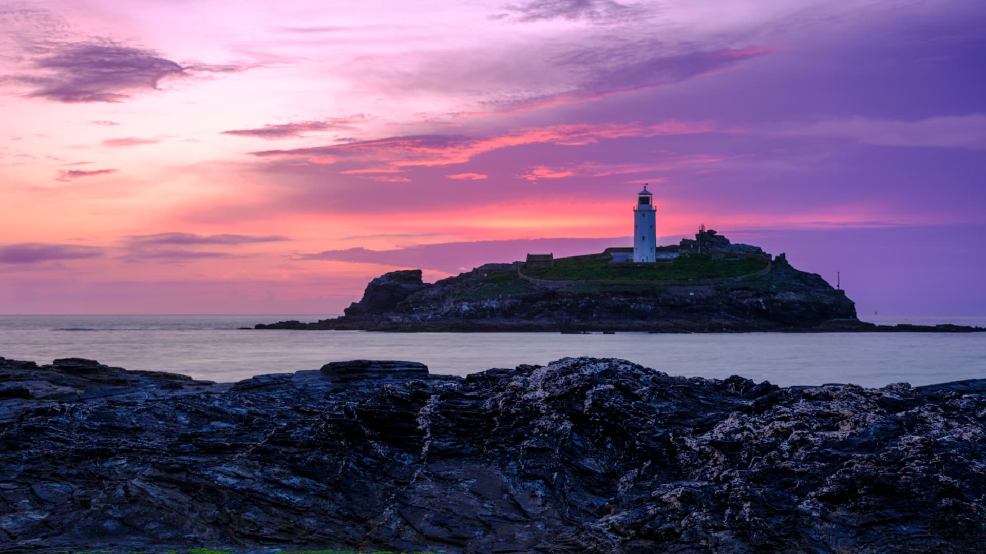 Lighthouse on rocky island at sunset with vibrant pink and purple sky over the sea