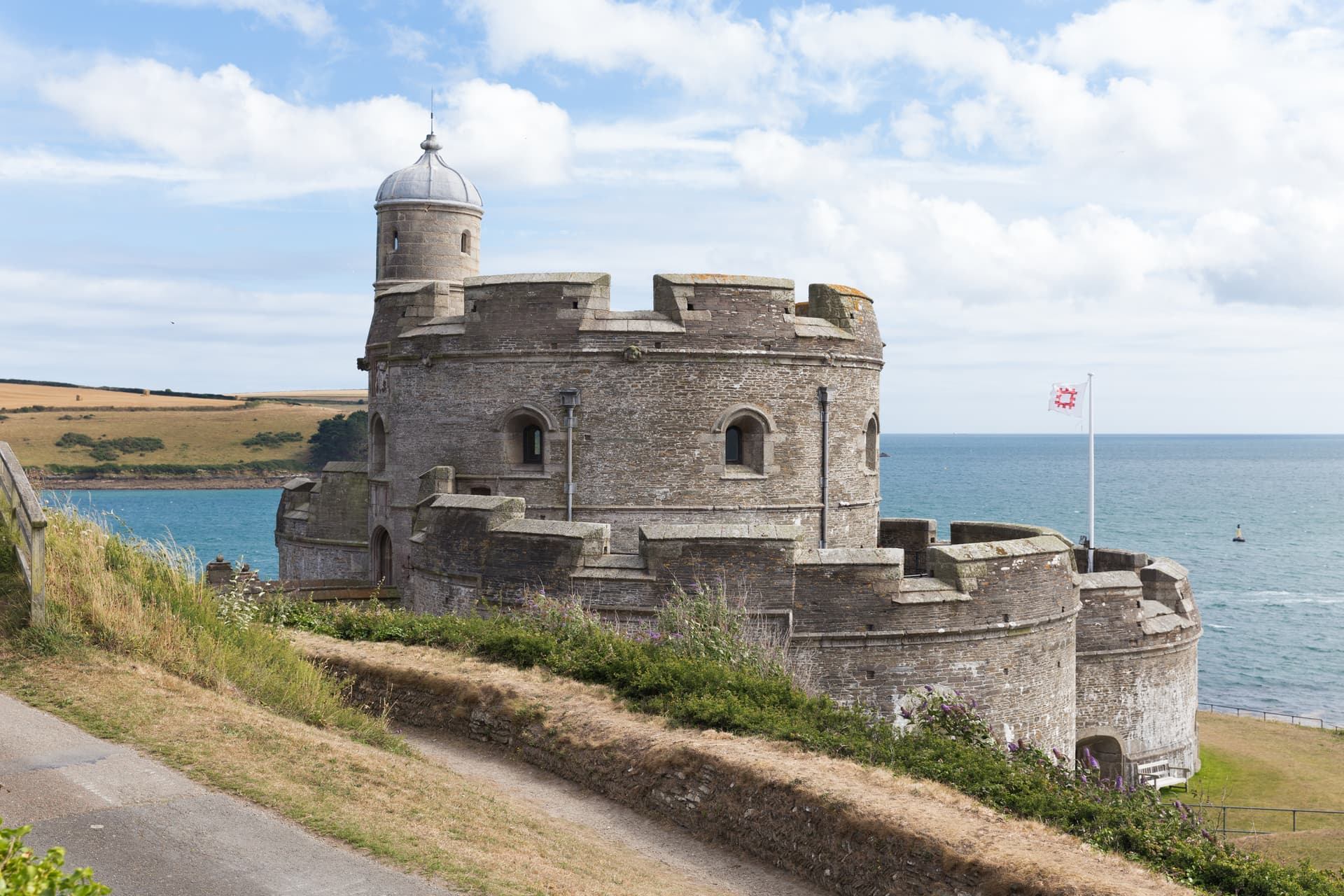 Stone coastal fort with a turret overlooking the sea and golden fields under a cloudy sky.
