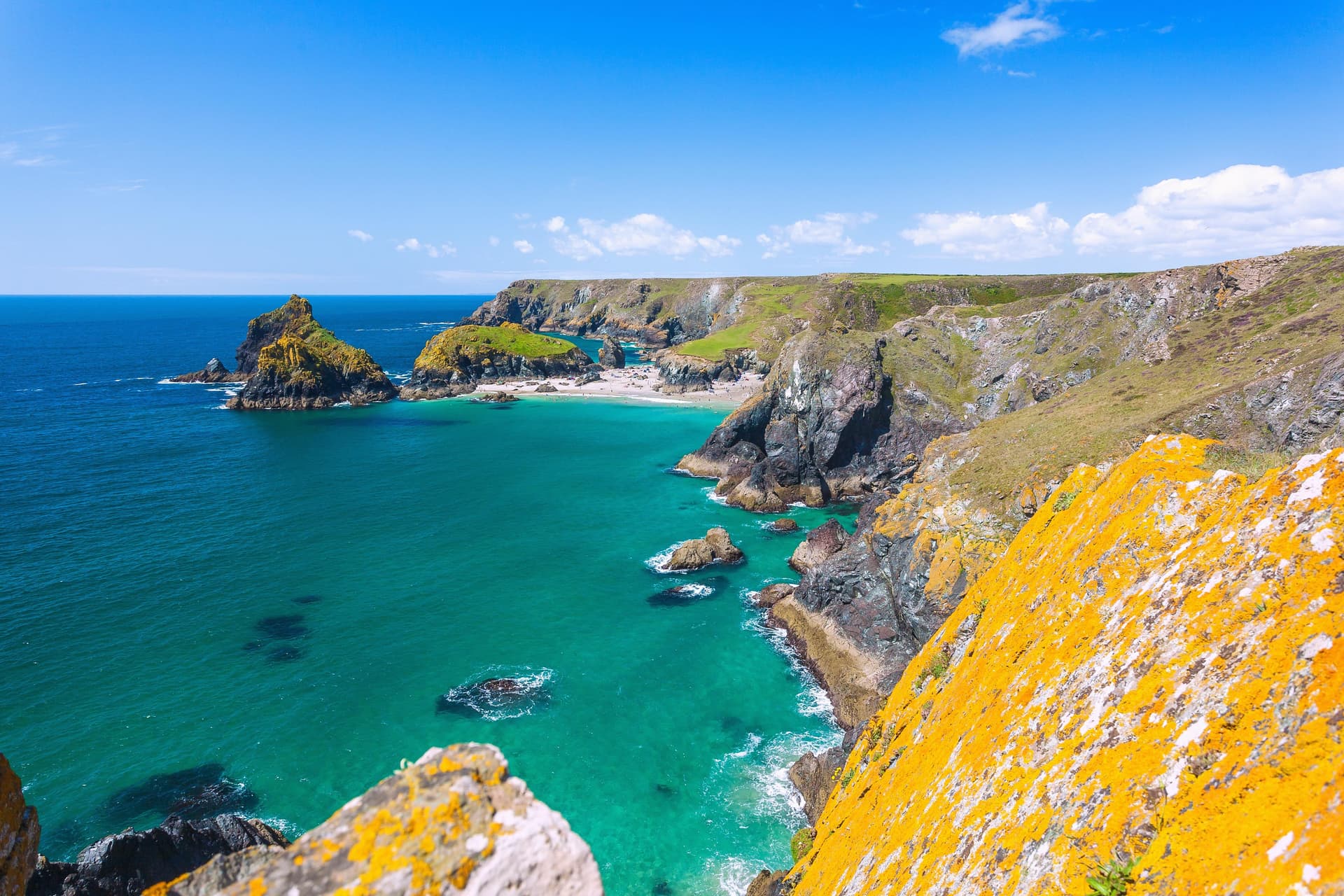 Coastal cliffs with bright orange lichen overlooking turquoise sea and a small sandy beach under blue sky.