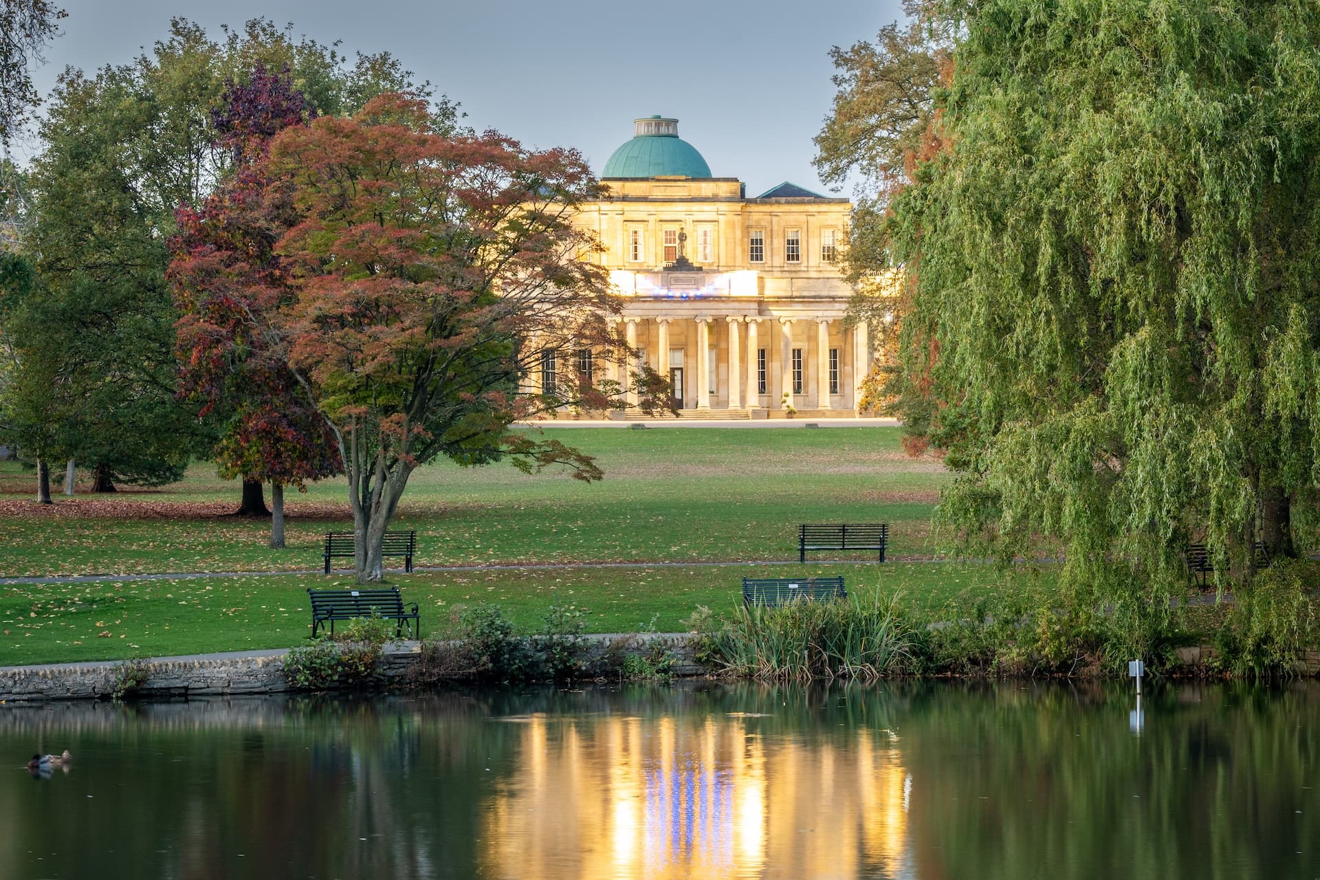 Neoclassical building with a dome reflected in a pond in a park with autumn trees.