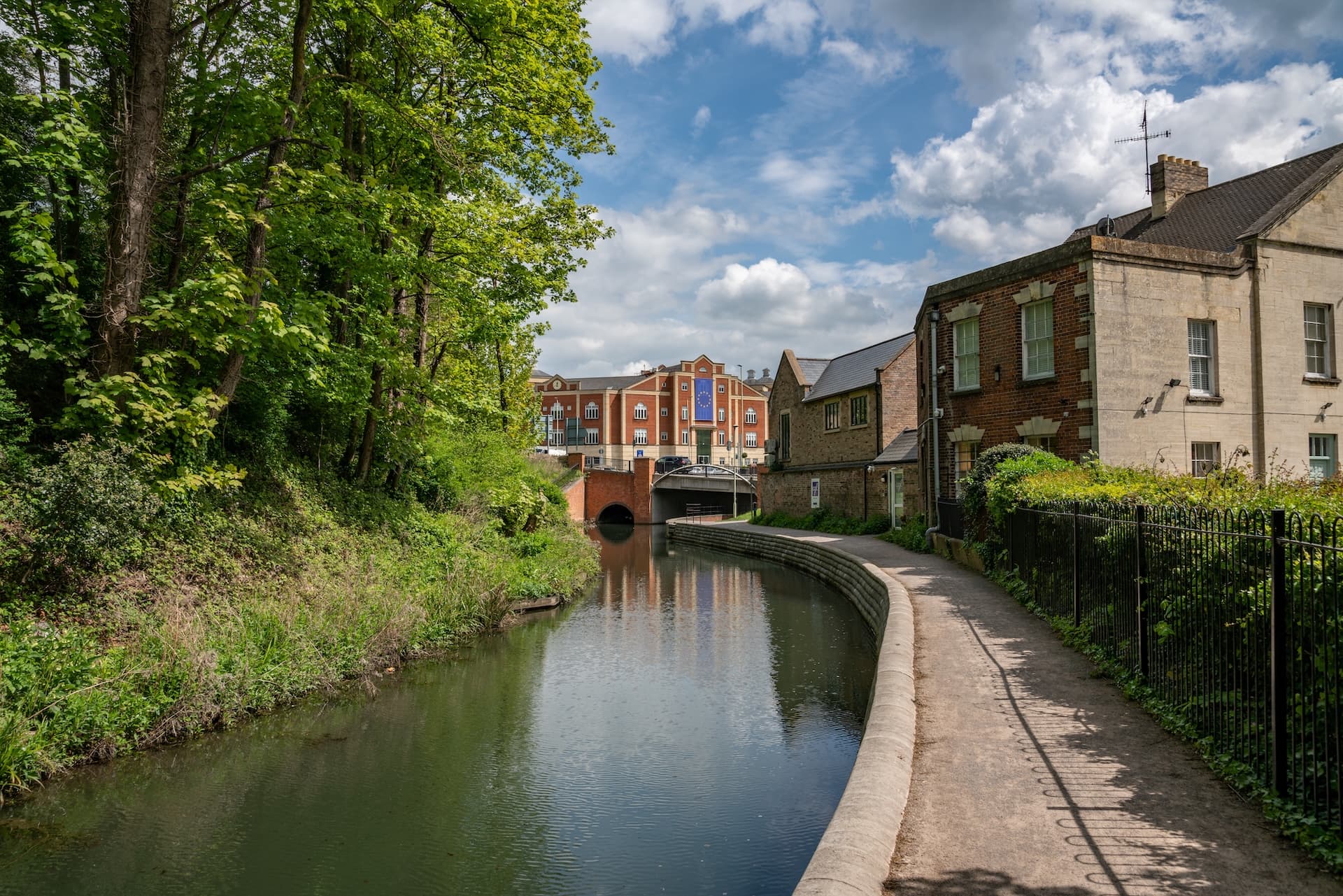 River canal walkway beside lush green trees leading toward a bridge and brick buildings under a cloudy sky.
