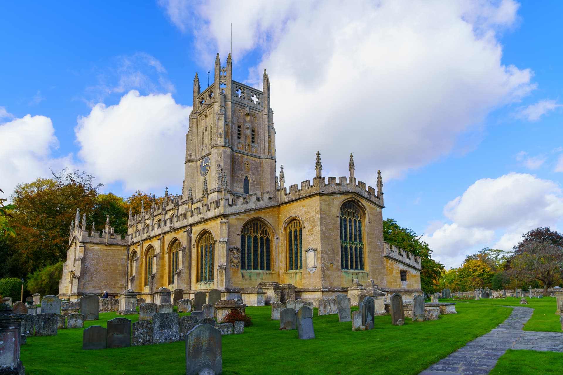 Stone church with tower and clock overlooking a green churchyard with gravestones in Fairford.