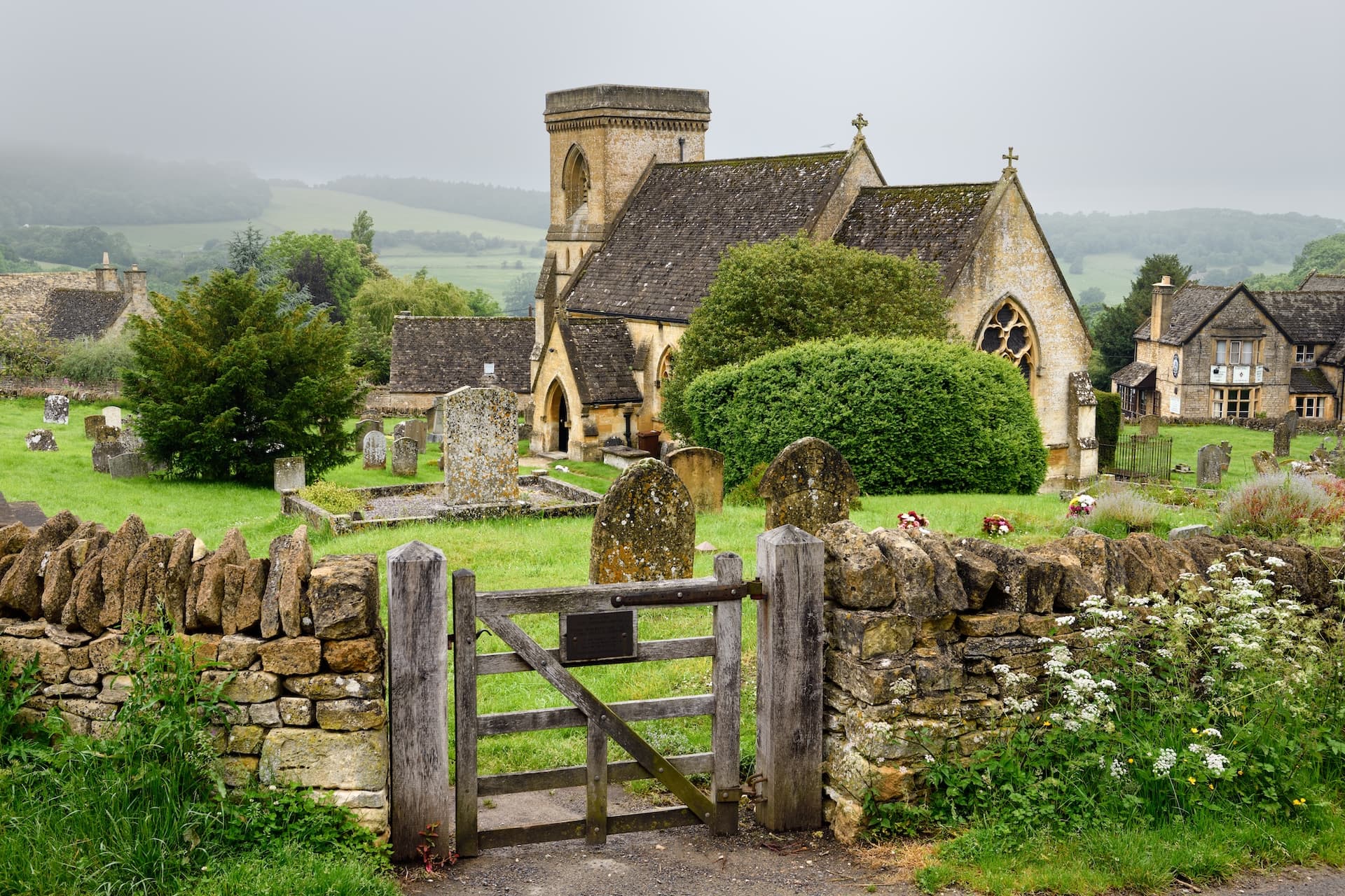 Stone churchyard with gravestones, dry stone wall, and wooden gate against foggy green hills.