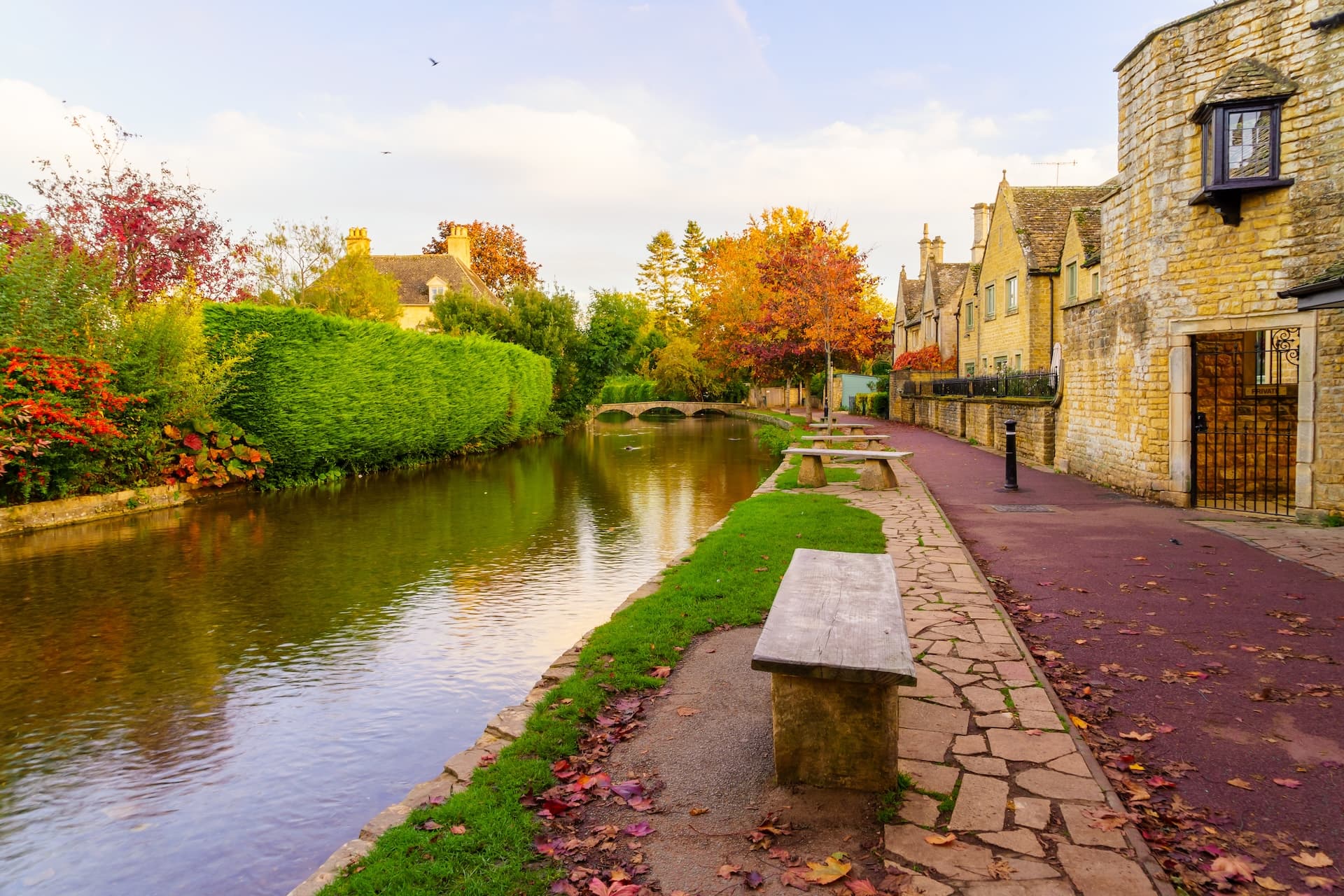 River walk in Bourton-on-the-Water with stone benches and autumn foliage by Cotswold stone buildings.