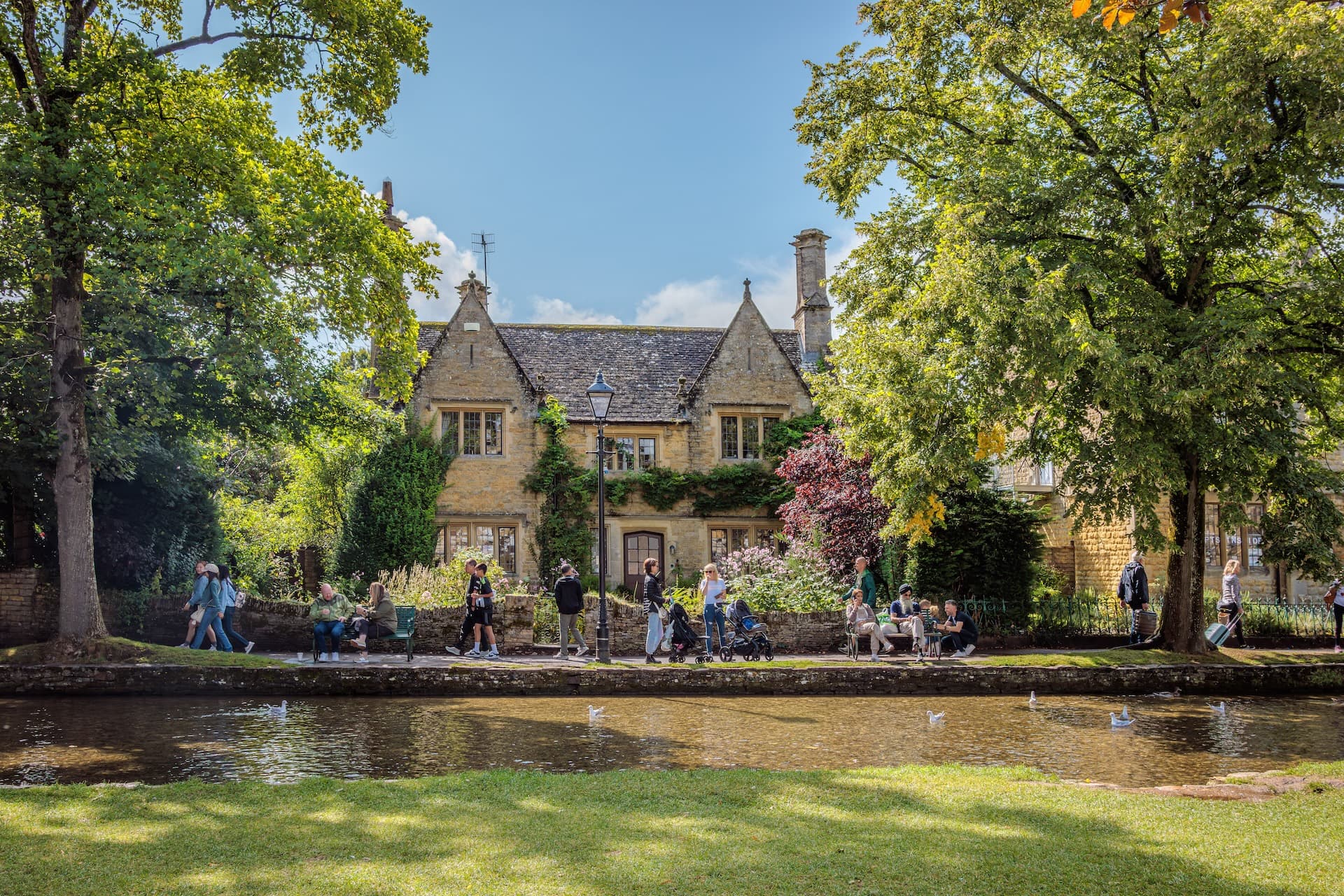 Cotswold stone building by river with people walking and sitting in Bourton-on-the-Water