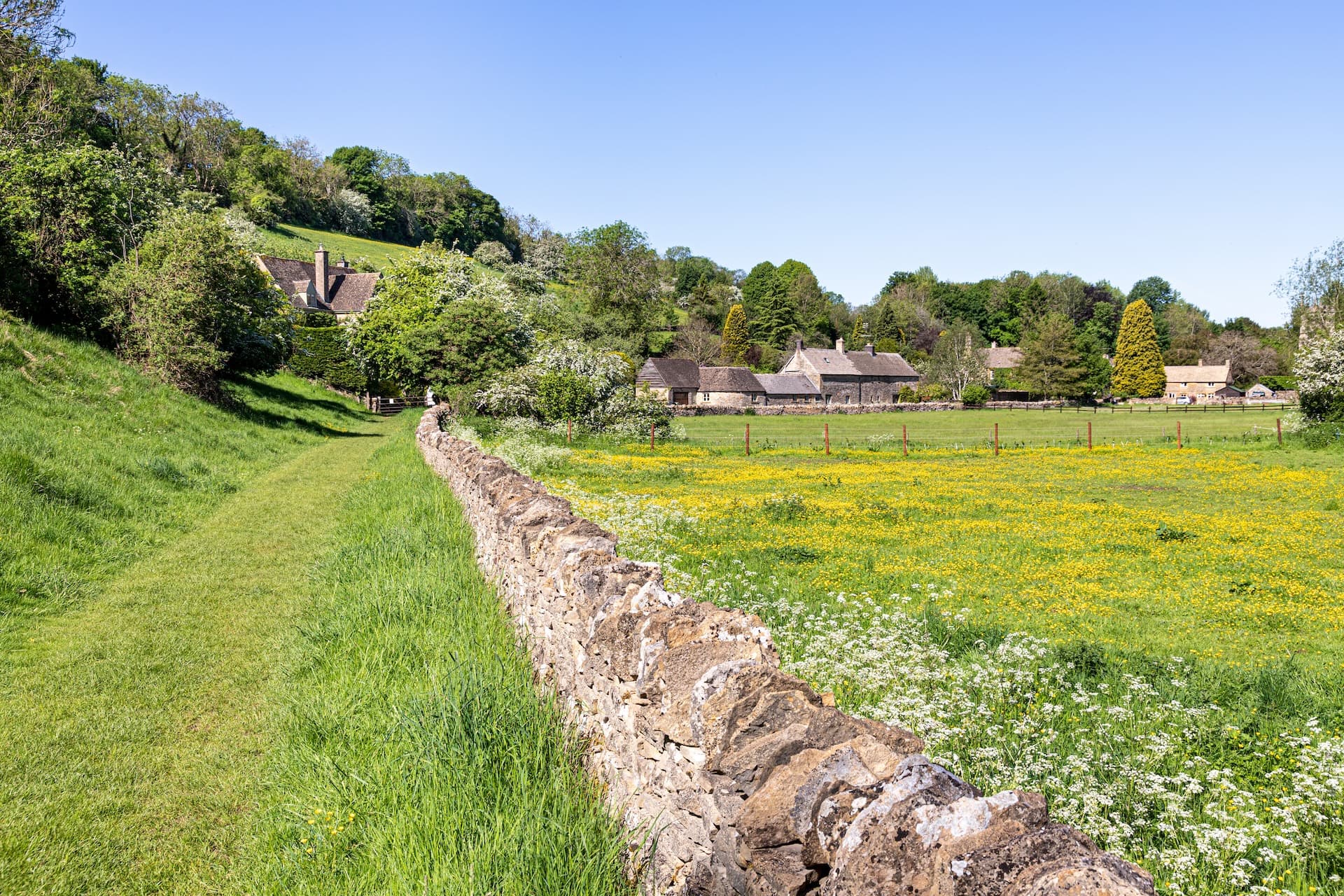Grassy footpath beside a dry stone wall leading toward stone cottages in Windrush Valley.