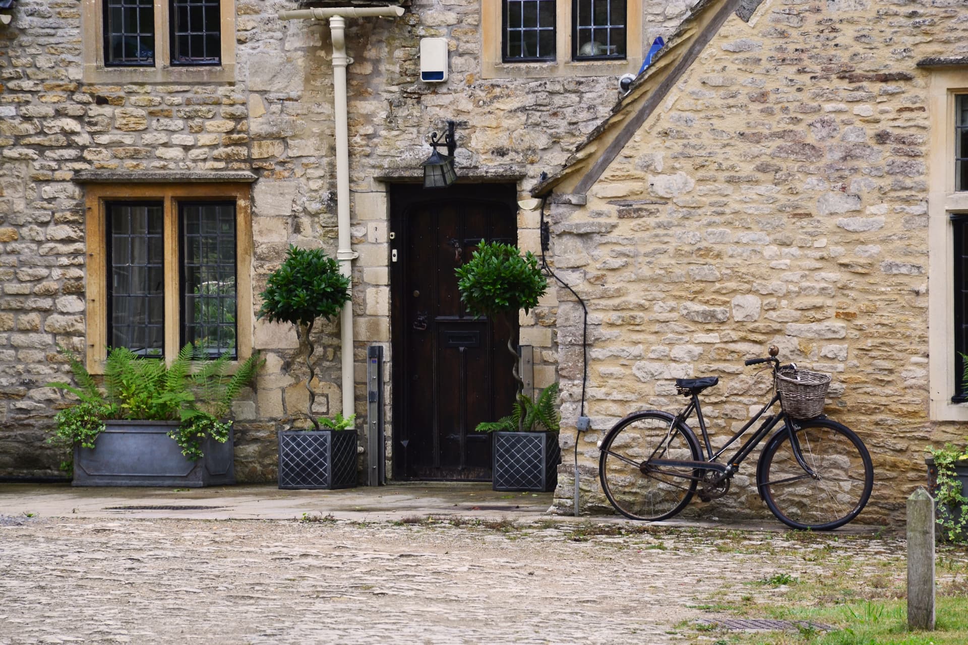 Bicycle parked by stone cottage entrance in the Cotswolds with ferns and potted trees.