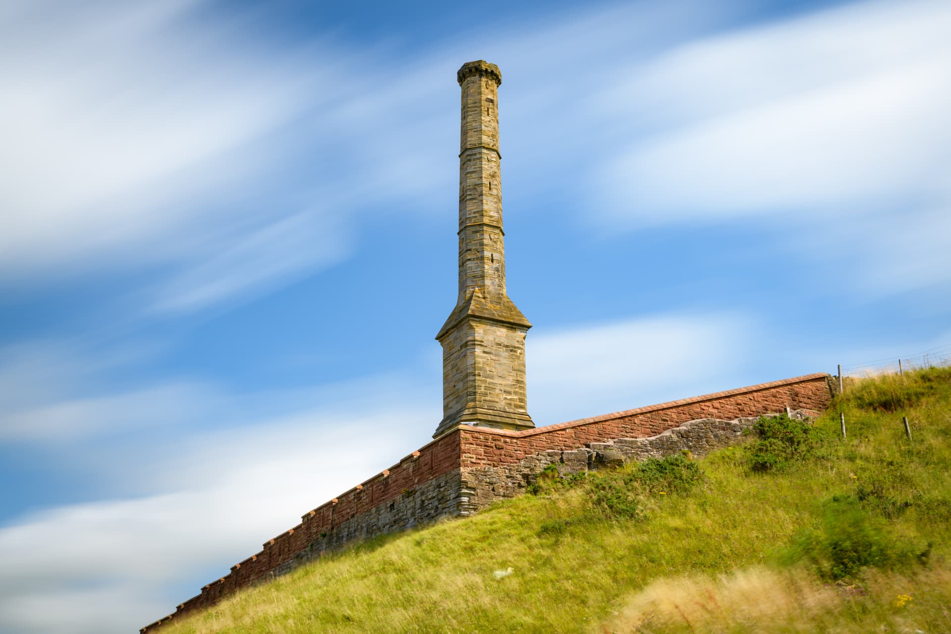 Stone chimney stack on a grassy hill next to a brick wall against a streaked blue sky.