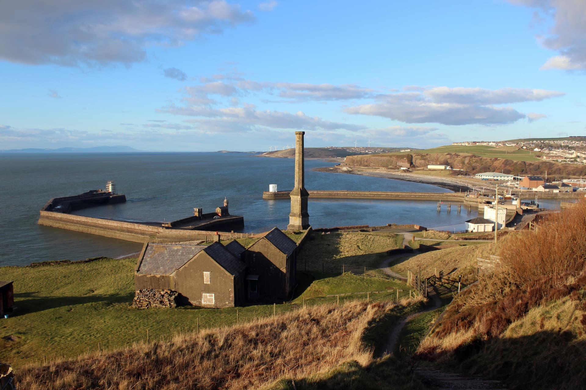 Stone chimney monument overlooking a harbor with breakwaters, sea, and coastal town.