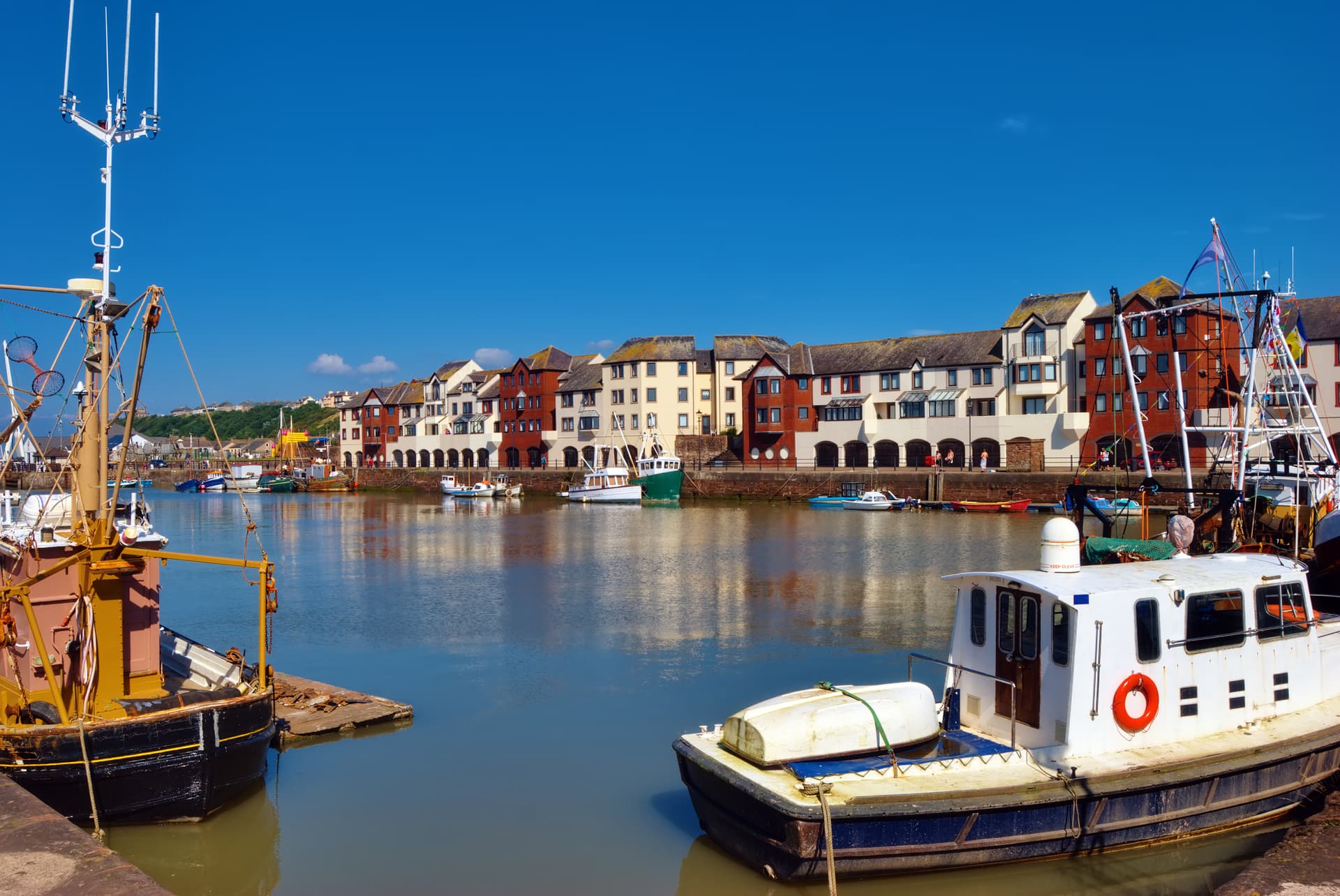 Boats moored in harbor with colorful waterfront buildings under a bright blue sky in Maryport.