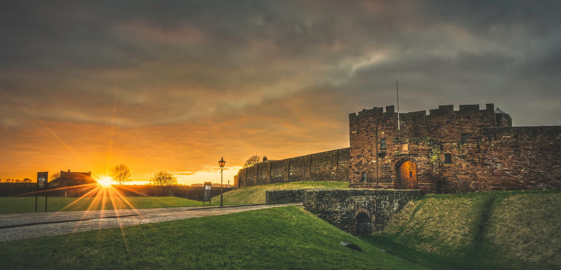 Carlisle Castle stone walls at sunset with sunburst over grassy moat and cobblestone path.