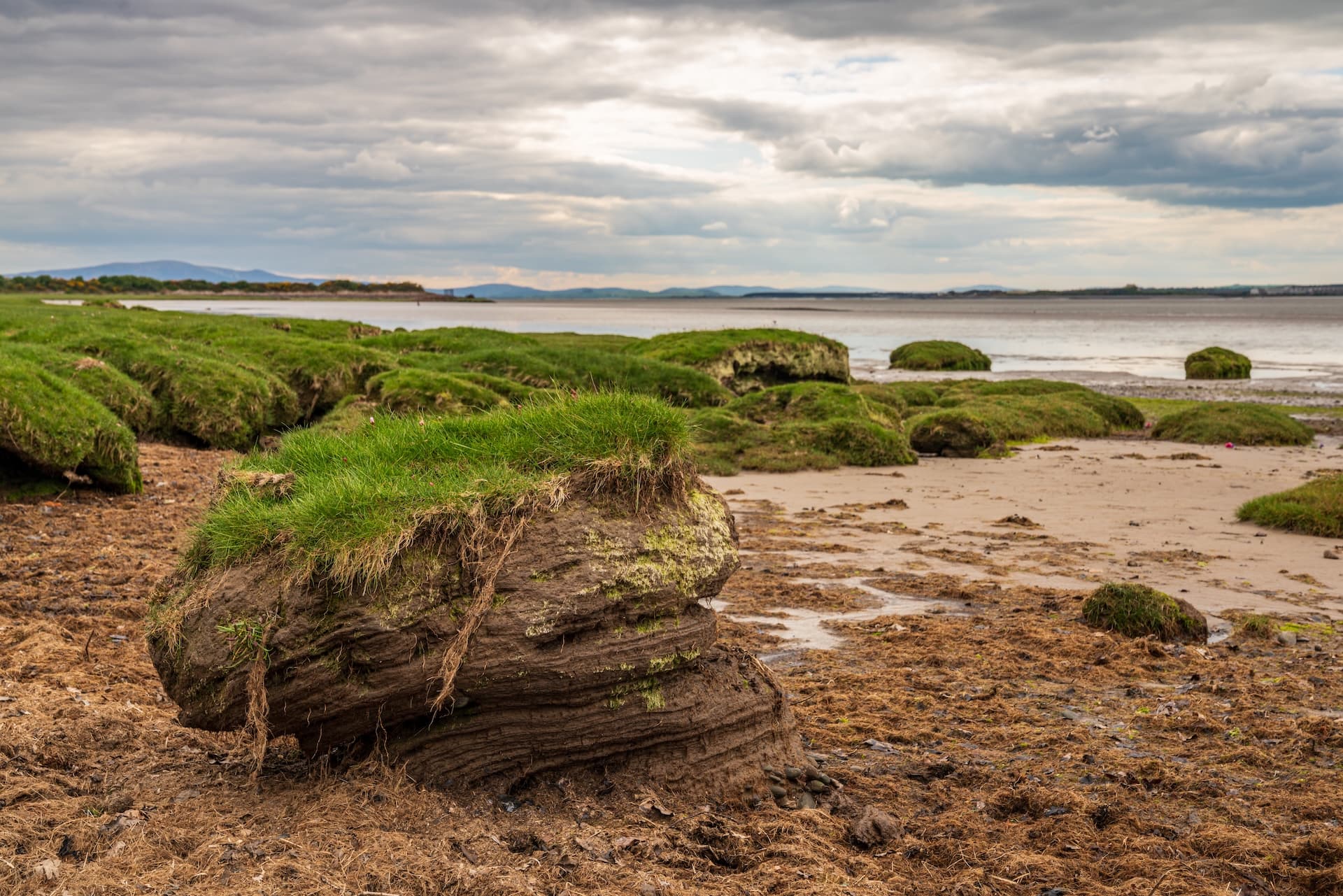 Eroded grassy mounds on muddy shore at Bowness-on-Solway under cloudy sky.