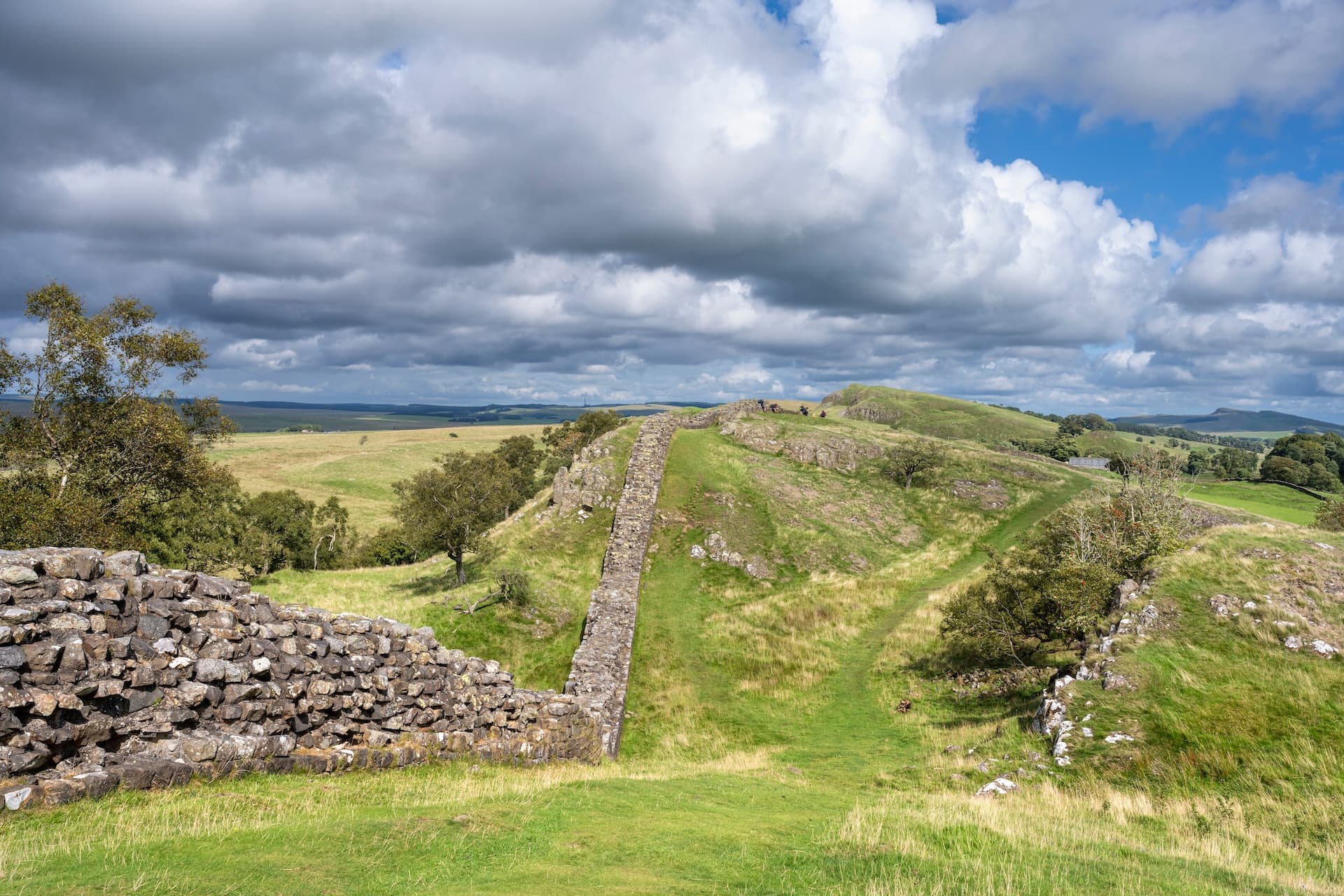 Stone wall ruins along green rolling hills under a dramatic cloudy sky at Hadrian's Wall.