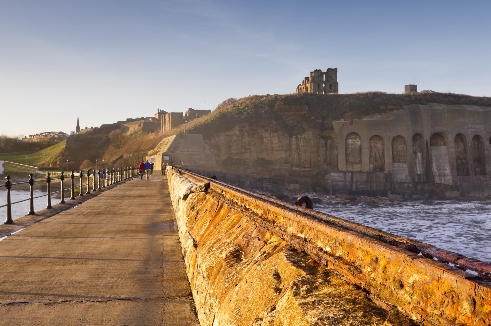 Walkway beside rusty sea wall towards Tynemouth Priory ruins on cliff top