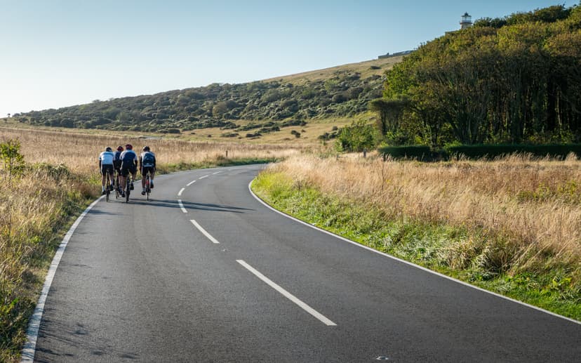 Cyclists riding on winding road past dry grassy hills toward a distant lighthouse.