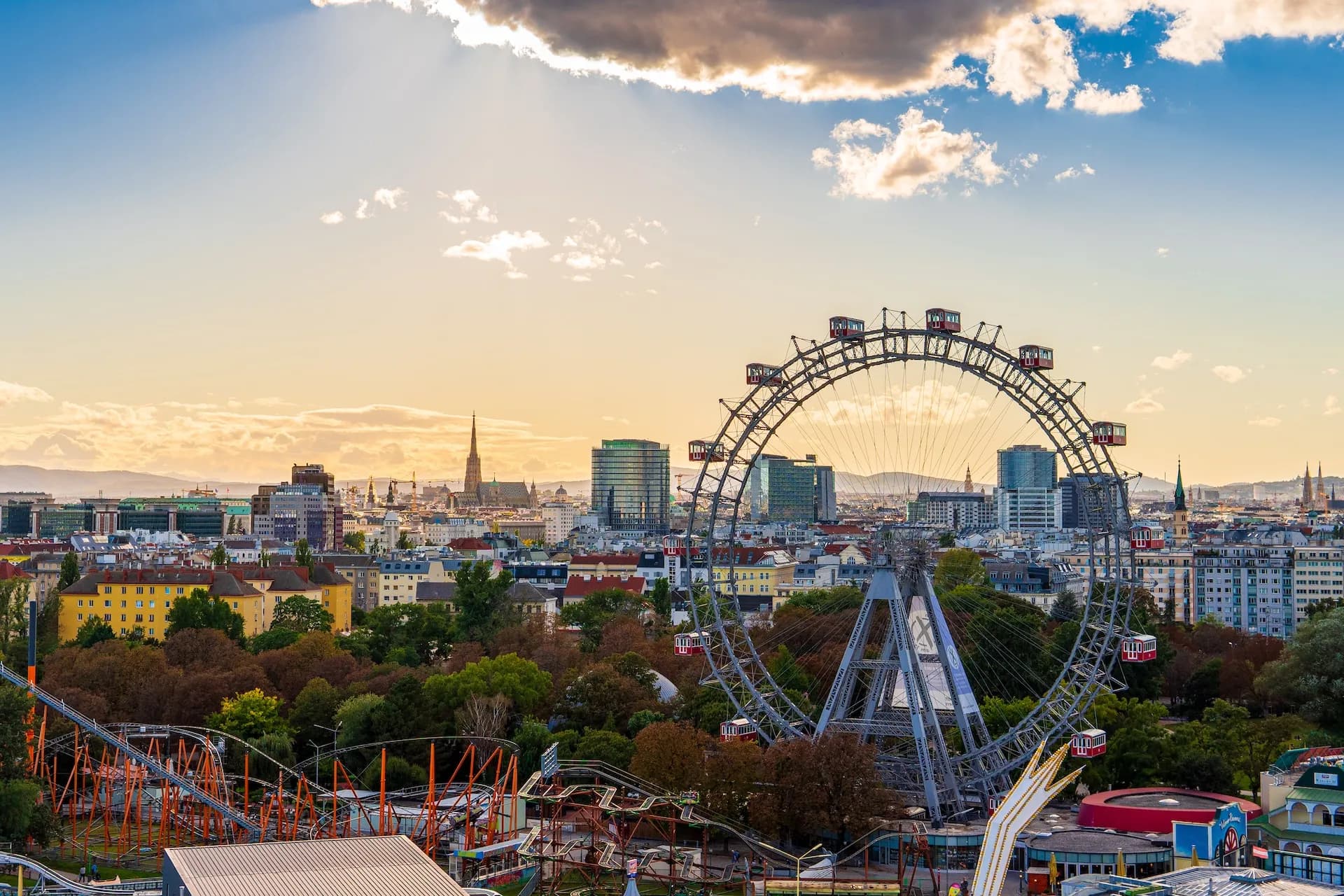 Giant Ferris wheel and roller coaster overlooking the Vienna cityscape at sunset.