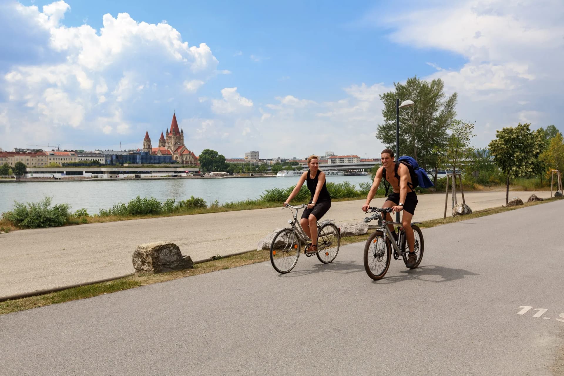 Two people cycling along a paved path next to a river with a city skyline featuring a red-roofed church.