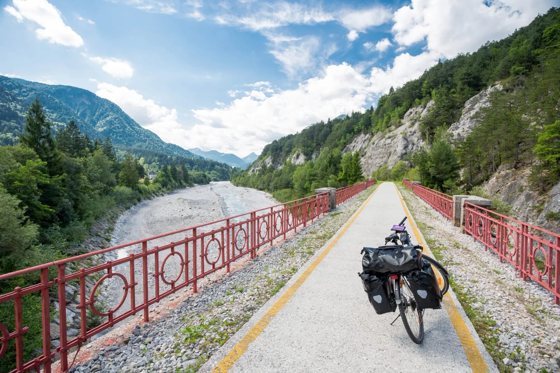 Bicycle touring on path beside riverbed with mountains and blue sky, Alpe Adria.