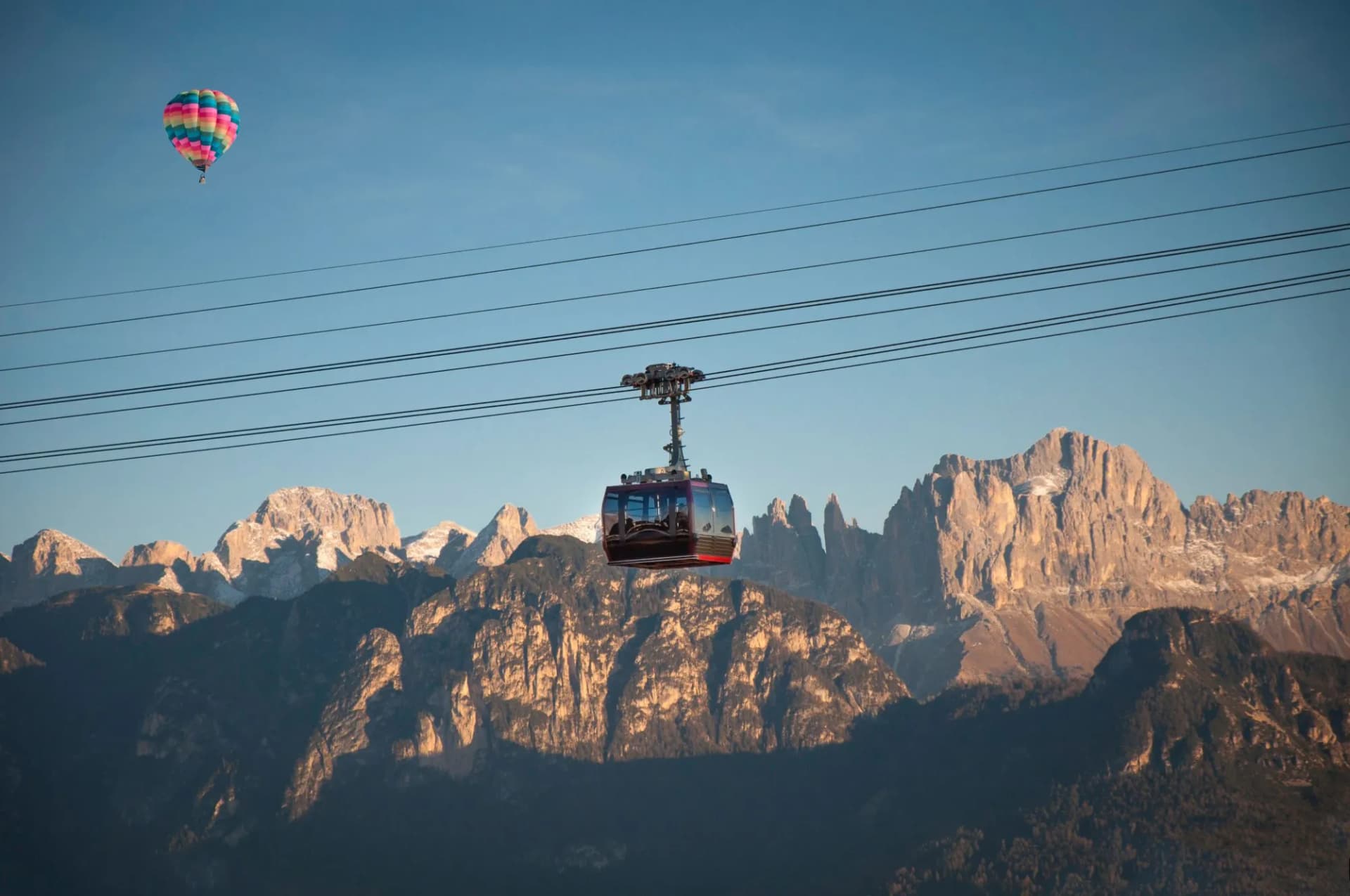 Cable car ascending over dark forested mountains with rocky peaks and a hot air balloon.