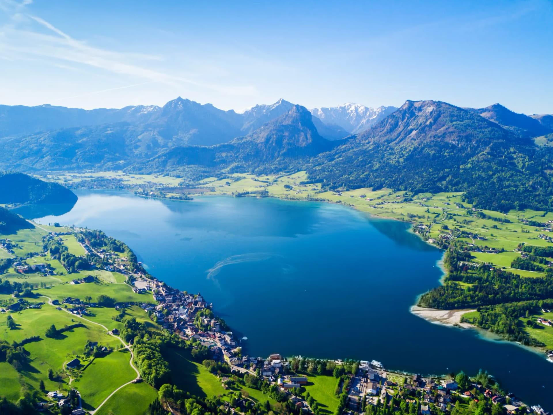 Aerial view of St. Wolfgang town on the shore of a deep blue lake with green hills and snow-capped mountains.