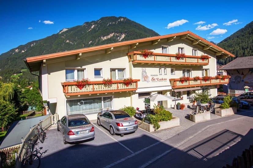 Pension St. Lukas building with balconies and flower boxes against a backdrop of forested mountains.