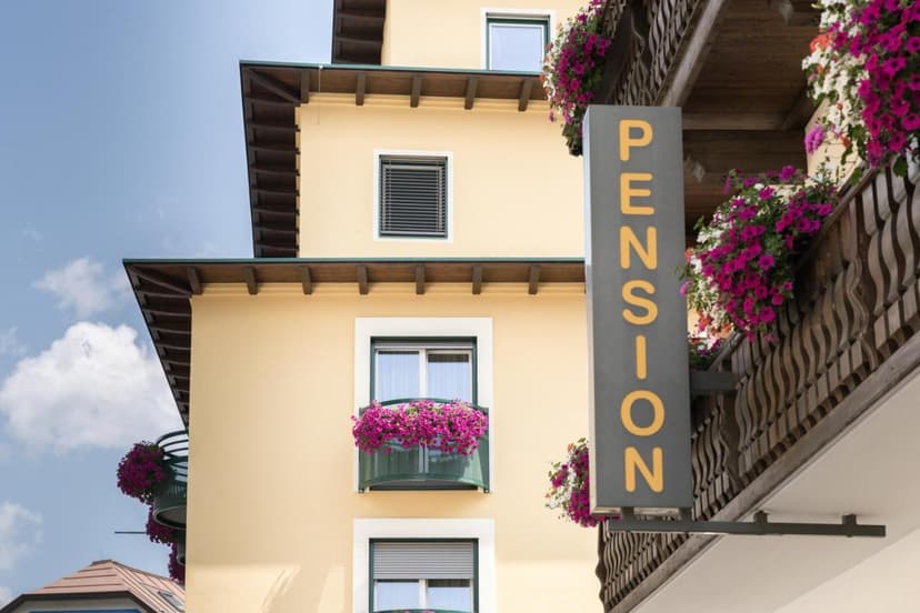 Pension sign next to yellow building with wooden balconies and bright pink flowers under blue sky.