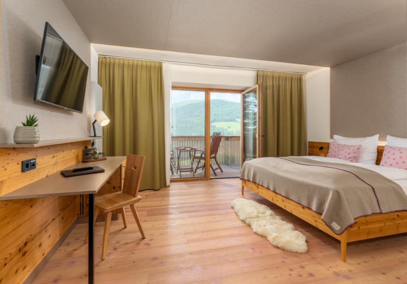Hotel room with wooden furniture, balcony view of green mountains, and light wood floors