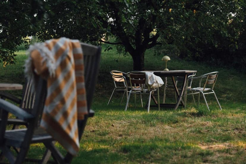 Outdoor seating area with wooden table, metal chairs, and striped blanket under a tree in Pension Obertrum am See.