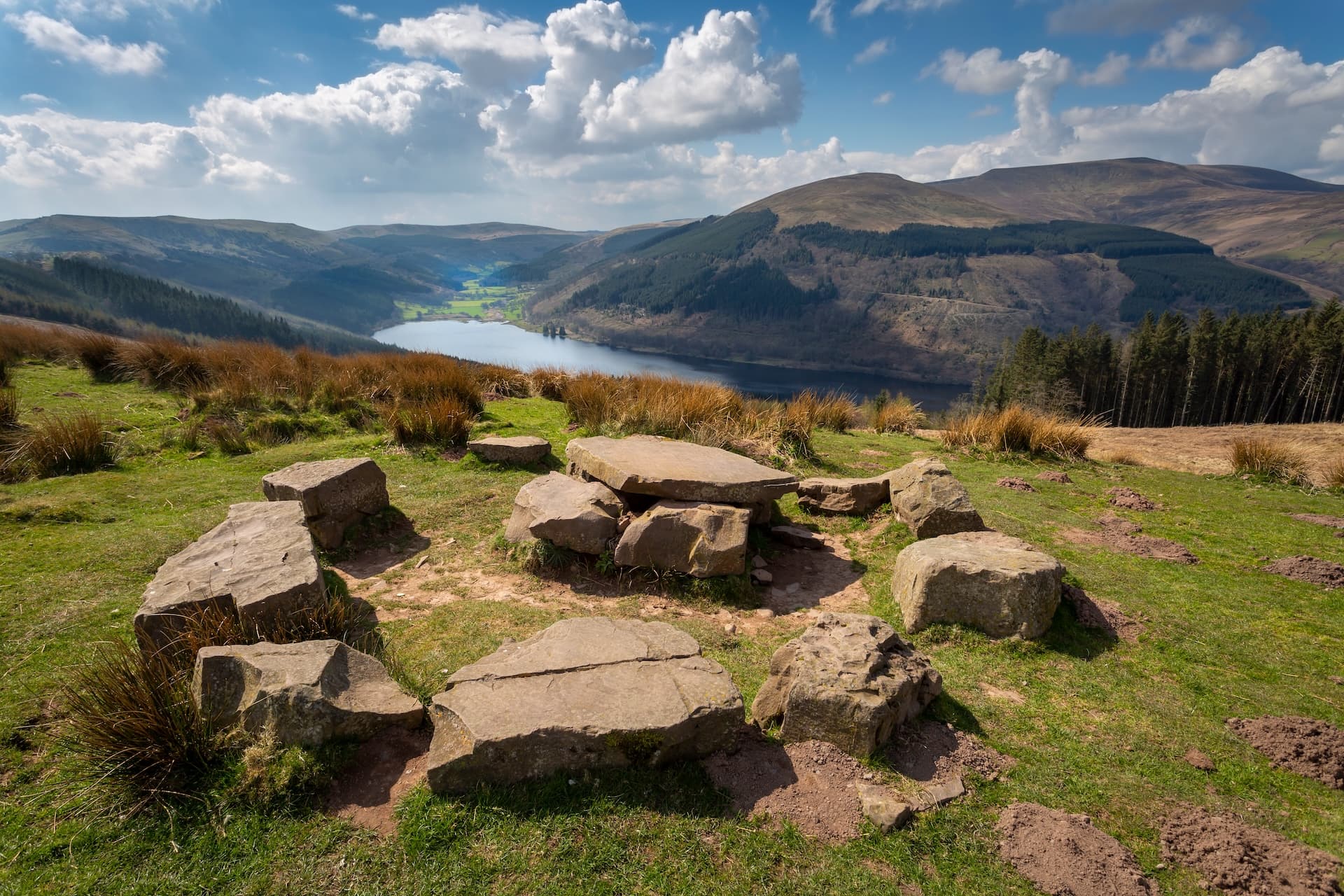 Stone seating area overlooking Talybont Reservoir and surrounding green hills under a blue sky.