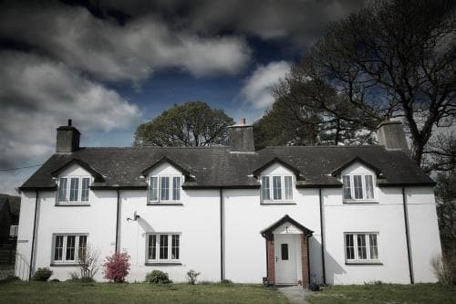 White cottage with dark roof under dramatic cloudy sky, trees in background