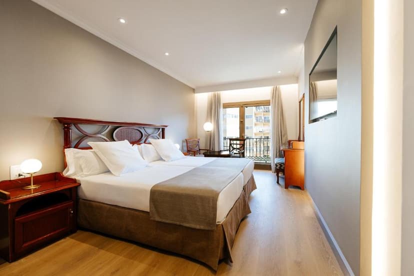 Hotel room with twin beds, wooden furniture, and balcony view of city buildings.