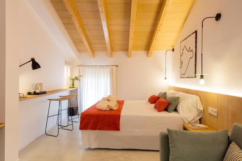 Bedroom with white walls, exposed wooden ceiling beams, and a bed with orange and green pillows.