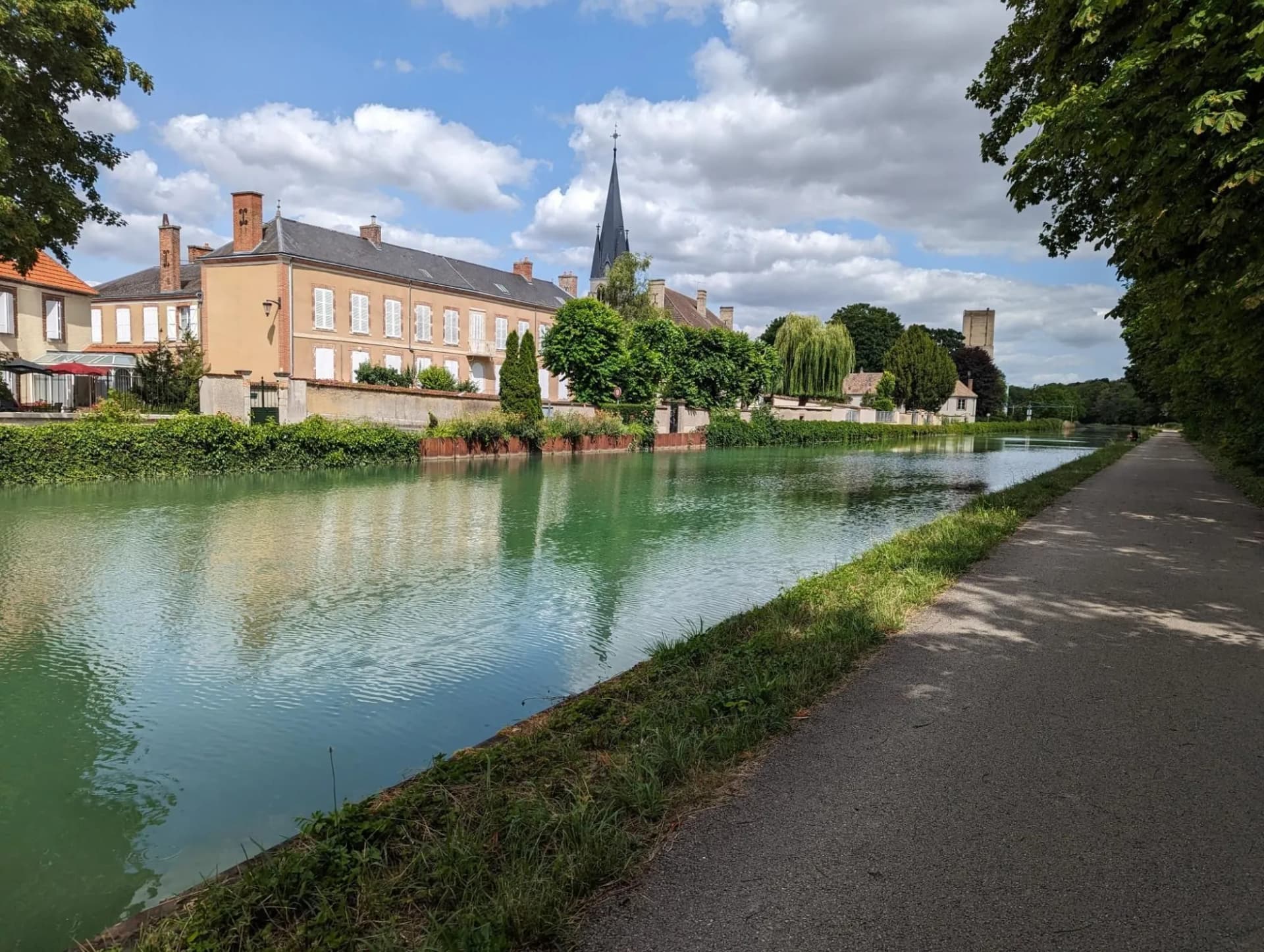 Canal path beside green water reflecting buildings and a church spire in Châlons-en-Champagne.