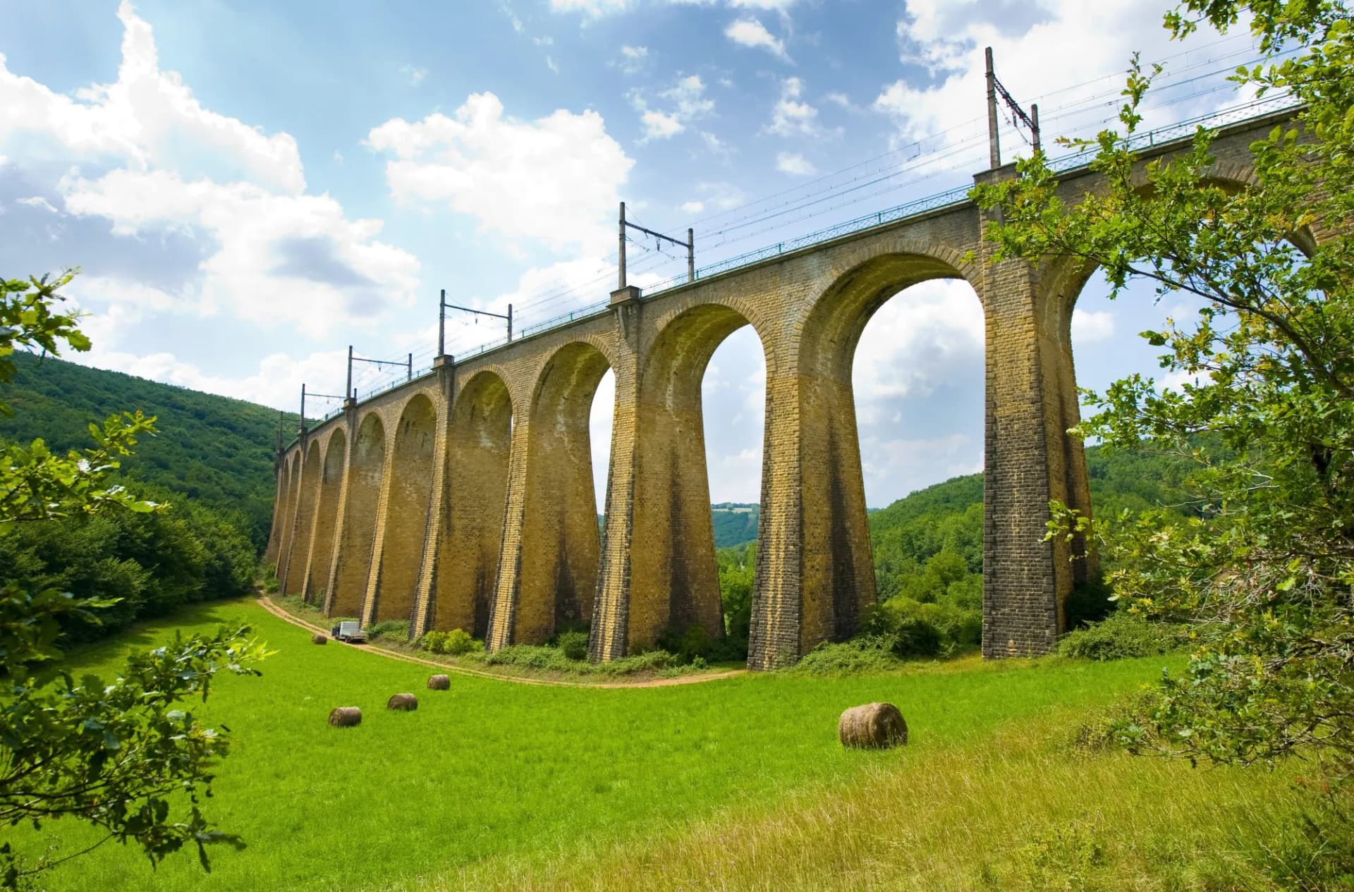 Stone arch viaduct spanning a green valley with hay bales under a partly cloudy sky in Souillac.